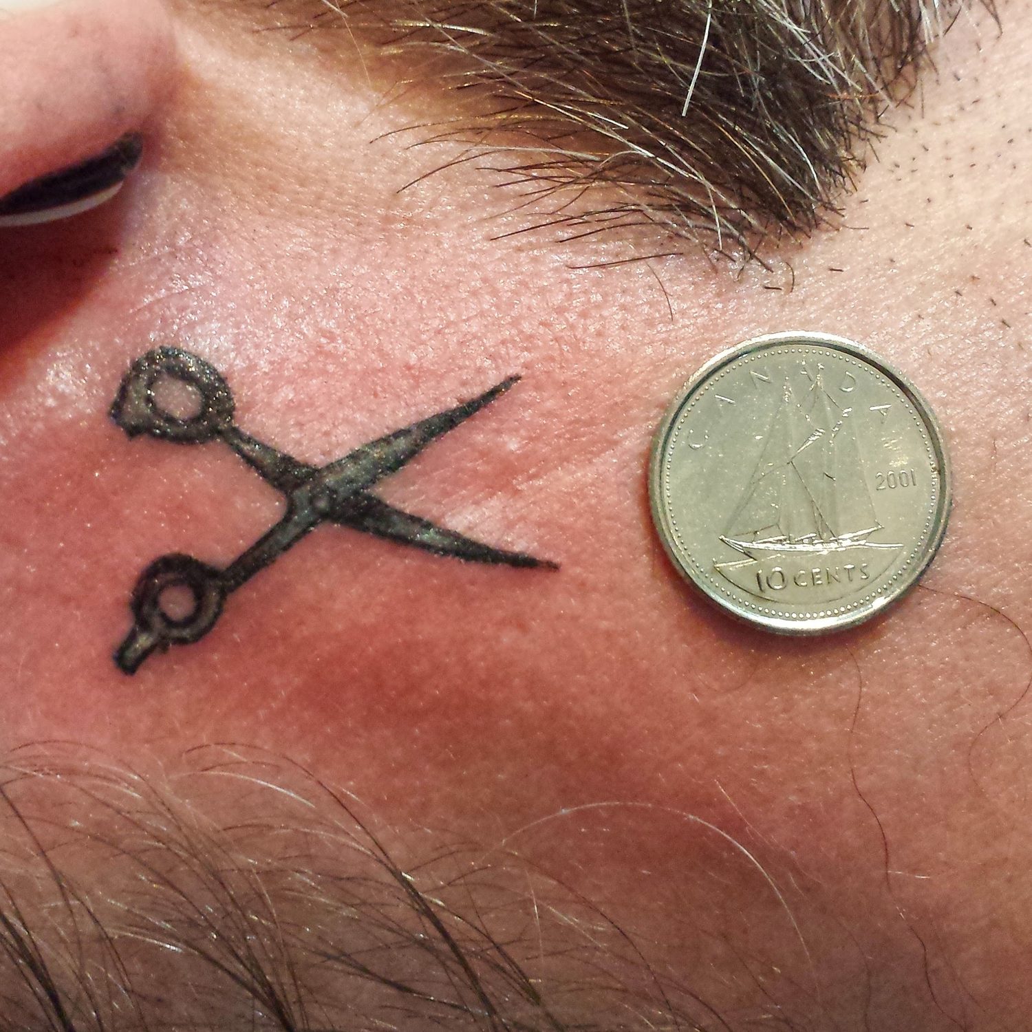 Tiny black scissors micro-tattoo by the ear, shown next to a dime for scale, Parksville BC